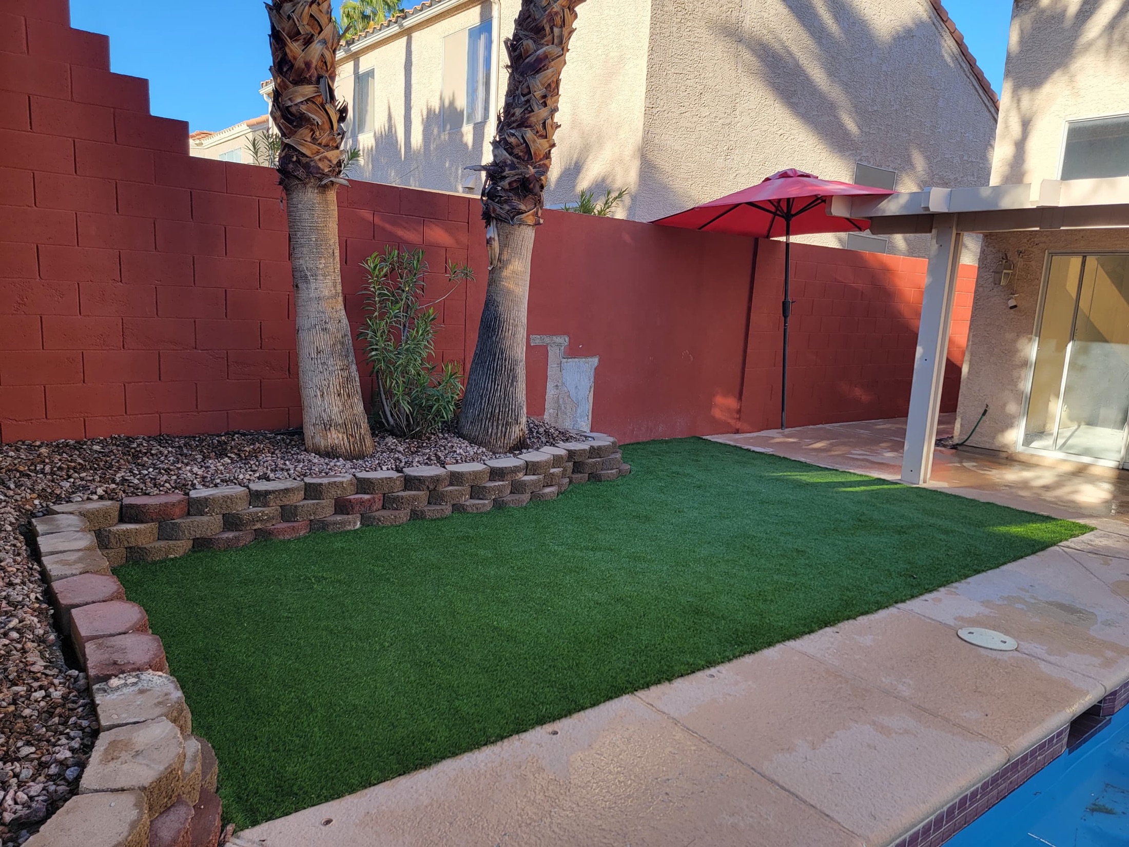 Artificial turf backyard with retaining block border, palm trees, rock, and patio umbrella